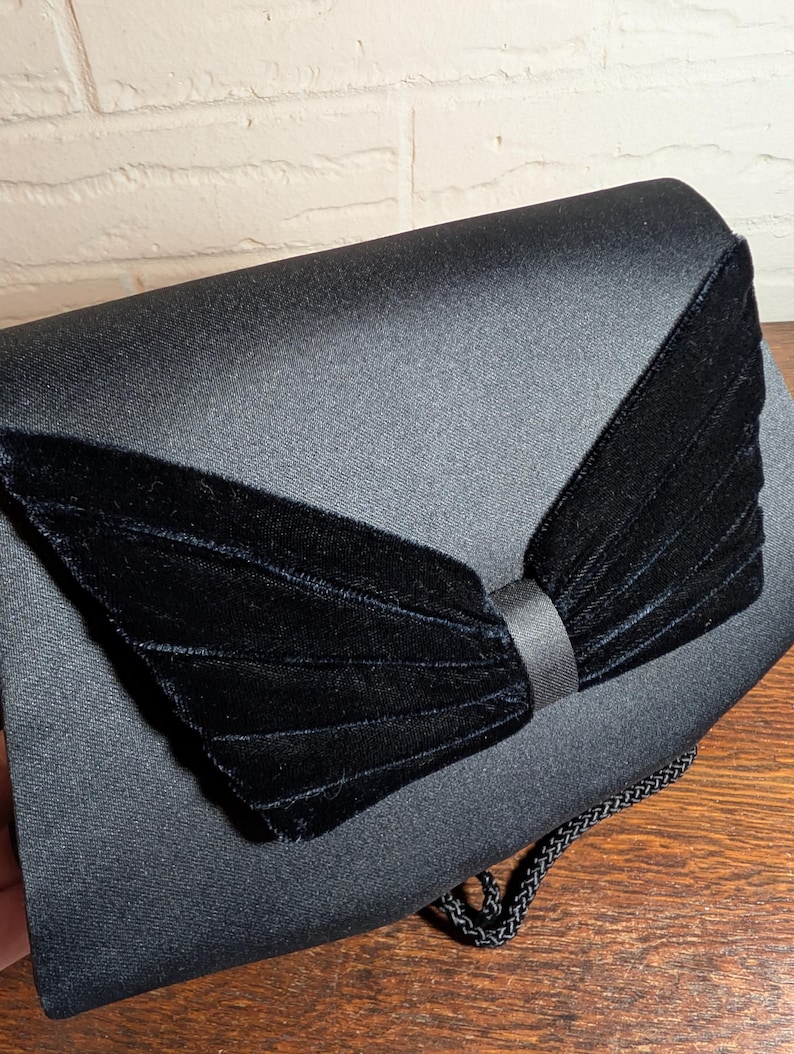 May include: A black satin clutch bag featuring a prominent, pleated black velvet bow detail on the front flap. The bag has a smooth, semi-glossy finish and includes a thin, braided black cord strap attached to the side for carrying or hanging.