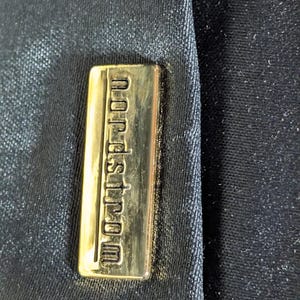 May include: A rectangular gold-toned metal plaque featuring the embossed text "nordstrom" in lowercase letters, securely attached to a dark black fabric surface with a subtle, fine-grained texture and a slight sheen across the material.