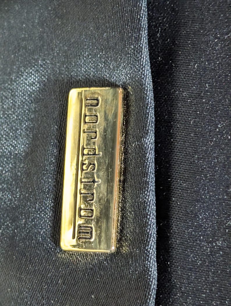 May include: A rectangular gold-toned metal plaque featuring the embossed text "nordstrom" in lowercase letters, securely attached to a dark black fabric surface with a subtle, fine-grained texture and a slight sheen across the material.