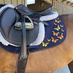 May include: A dark brown leather horse saddle with a navy blue saddle pad decorated with orange and yellow butterfly appliqu&eacute;s. The saddle has a white fleece lining and silver stirrups. The saddle is on a brown horse.