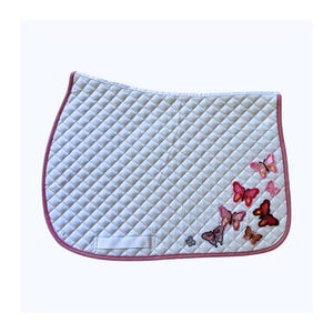 May include: White quilted saddle pad with a pink trim and butterfly embroidery. The pad has a diamond pattern and a small white strap. The butterflies are in shades of pink and red.