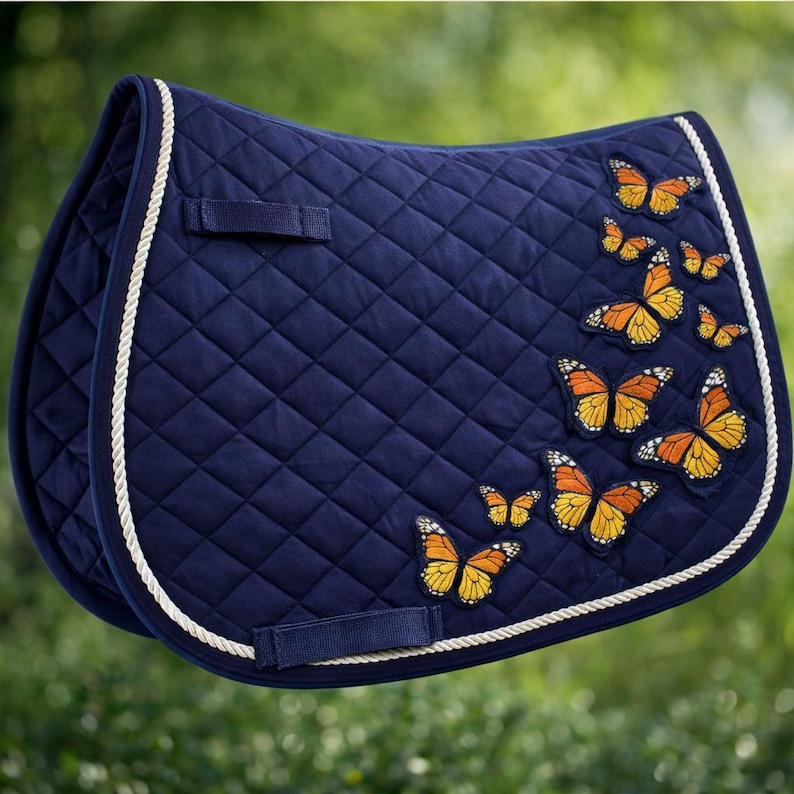 May include: A navy blue quilted saddle pad with a white rope trim. The pad is decorated with embroidered orange and yellow butterflies. The pad has two straps and a loop for the saddle.