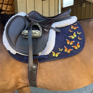 May include: A dark brown leather saddle with a navy blue quilted pad featuring orange and yellow butterfly appliqu&eacute;s. The saddle has a white sheepskin lining and silver stirrups. The saddle is on a brown horse.