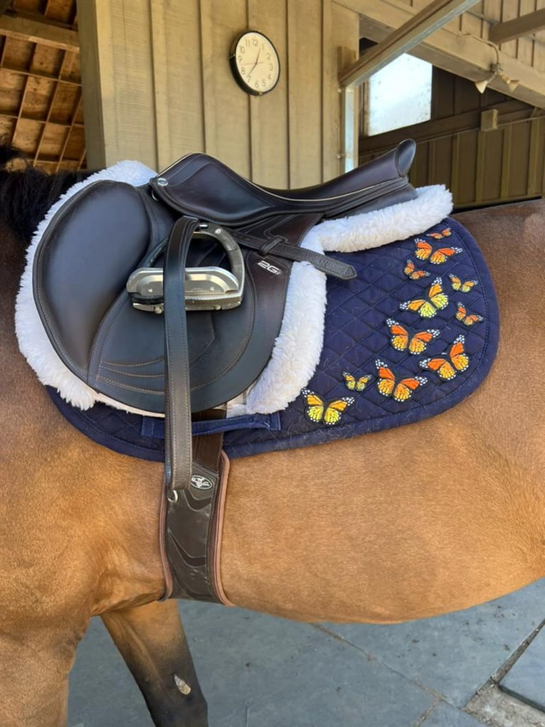 May include: A dark brown leather saddle with a navy blue quilted pad featuring orange and yellow butterfly appliqu&eacute;s. The saddle has a white sheepskin lining and silver stirrups. The saddle is on a brown horse.