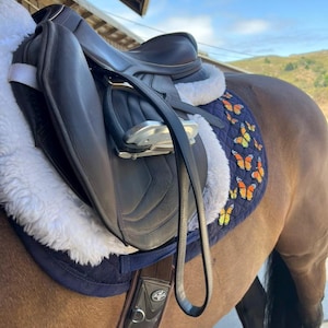 May include: A black leather horse saddle with a white sheepskin lining and a navy blue pad featuring orange and yellow butterfly designs. The saddle is on a brown horse, with a black leather strap.