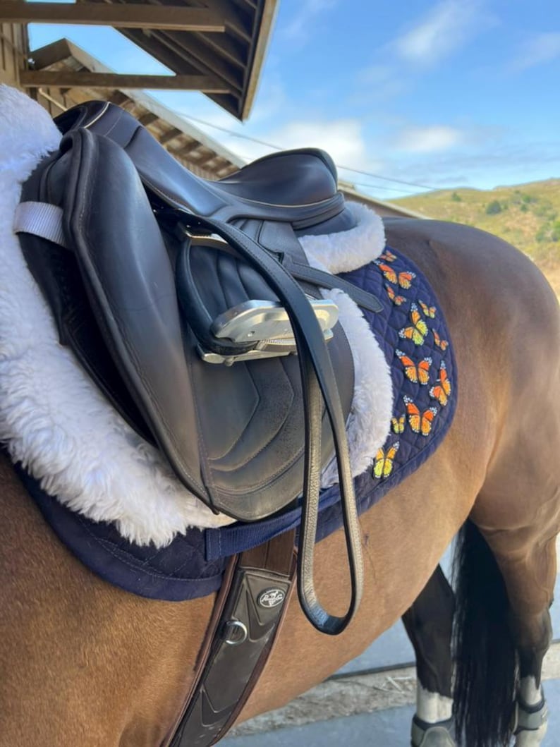 May include: A black leather horse saddle with a white sheepskin lining and a navy blue pad featuring orange and yellow butterfly designs. The saddle is on a brown horse, with a black leather strap.