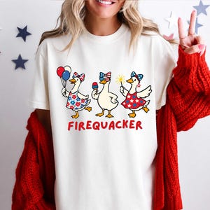 May include: Cream-colored t-shirt featuring three cartoon geese in patriotic outfits. The geese are holding balloons, ice cream, and a sparkler. The word "FIREQUACKER" is printed in red below the geese.