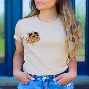 May include: Beige t-shirt with a retro "Summer 2023" design featuring palm trees and a sun graphic. The design is in a triangle shape with a vintage color palette. The shirt is paired with blue jeans.