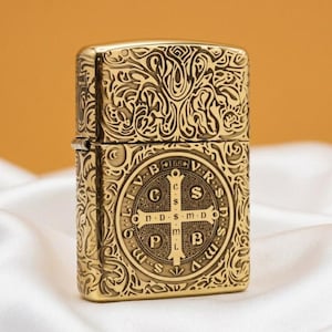 May include: A gold-coloured metal lighter with detailed engraved patterns. The design features a cross and Latin text. The lighter has a hinged lid and a rectangular shape. The overall aesthetic is ornate and detailed.
