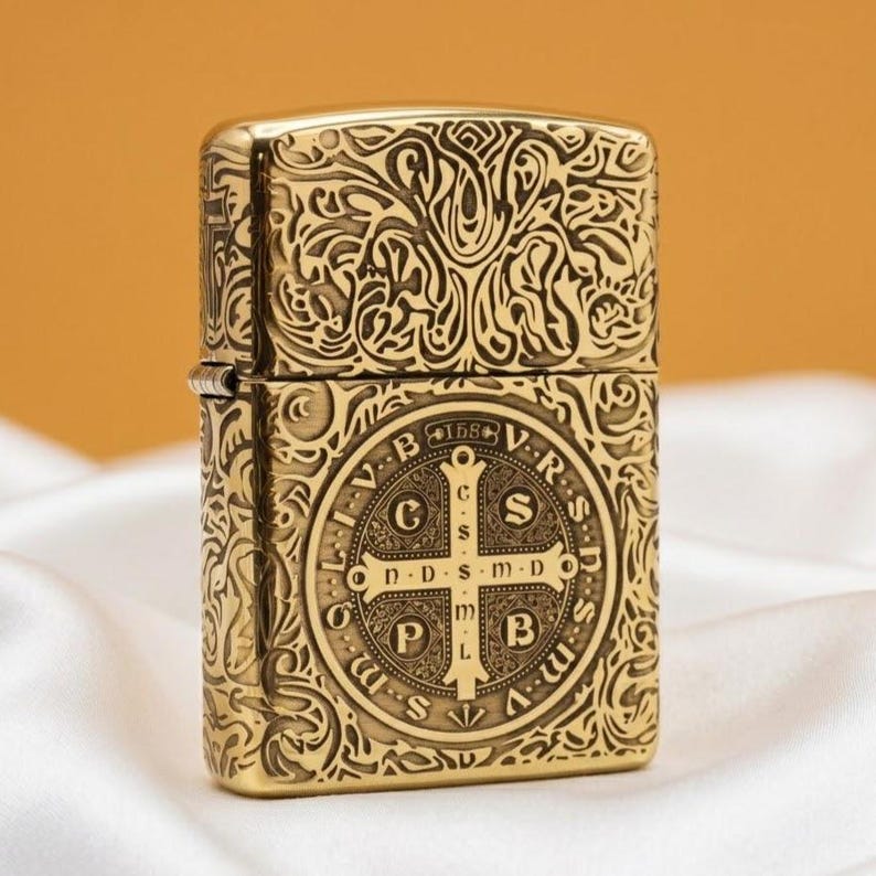 May include: A gold-coloured metal lighter with detailed engraved patterns. The design features a cross and Latin text. The lighter has a hinged lid and a rectangular shape. The overall aesthetic is ornate and detailed.