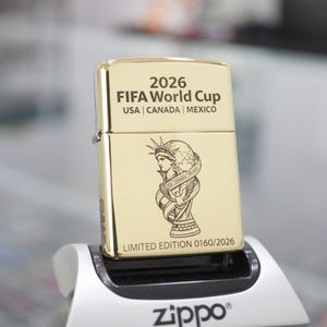 以下が含まれることがあります： 上部に「2026 FIFA World Cup USA | CANADA | MEXICO」と刻印されたゴールドトーンの金属製ライター。中央には自由の女神像とトロフィーのイラストが描かれ、下部には「LIMITED EDITION 0160/2026」と刻印されています。