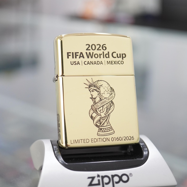 以下が含まれることがあります： 上部に「2026 FIFA World Cup USA | CANADA | MEXICO」と刻印されたゴールドトーンの金属製ライター。中央には自由の女神像とトロフィーのイラストが描かれ、下部には「LIMITED EDITION 0160/2026」と刻印されています。