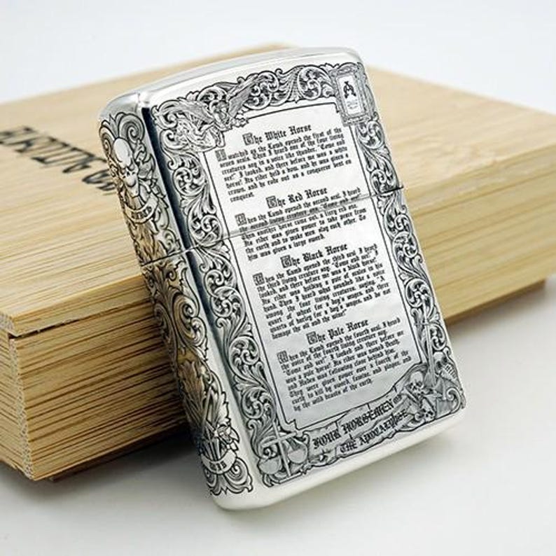 May include: Silver-colored metal lighter with intricate engraved detailing. The lighter features ornate scrollwork, a skull, and text. The text includes the words "The White Horse", "The Red Horse", "The Black Horse", and "The Pale Horse".