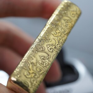Pode incluir: Um isqueiro dourado com um padr&atilde;o ornamental e rodopiante gravado em sua superf&iacute;cie. O design apresenta um motivo repetitivo de nuvens e curvas estilizadas, criando uma apar&ecirc;ncia texturizada. A forma retangular do isqueiro &eacute; vis&iacute;vel, com um acabamento polido.