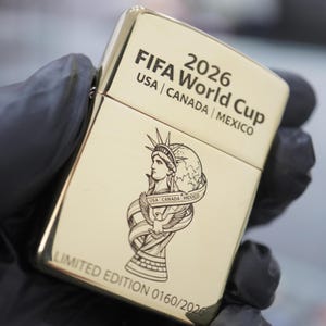 以下が含まれることがあります： 上部に「2026 FIFA World Cup USA | CANADA | MEXICO」の文字が刻まれたゴールドカラーの金属製ライター。文字の下にはリボンをまとった自由の女神像の彫刻が施されている。下部には「LIMITED EDITION 0160/2026」と記されている。