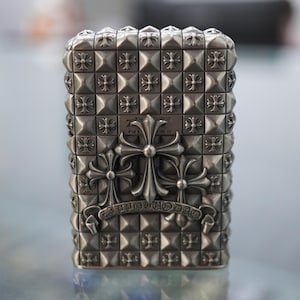 May include: A silver metal lighter with a studded design and cross emblems. The lighter has a rectangular form, covered in small, square studs. Three elaborate crosses are centered on the front, with a banner beneath them.
