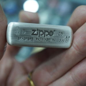 K&ouml;nnte beinhalten: Ein Feuerzeug der Marke Zippo aus geb&uuml;rstetem Metall mit dem Markennamen und der Gravur "25" auf der Oberfl&auml;che. Der Text "BRADFORD, PA. MADE IN U.S.A." ist ebenfalls sichtbar. Das Feuerzeug wird von einer Person gehalten.