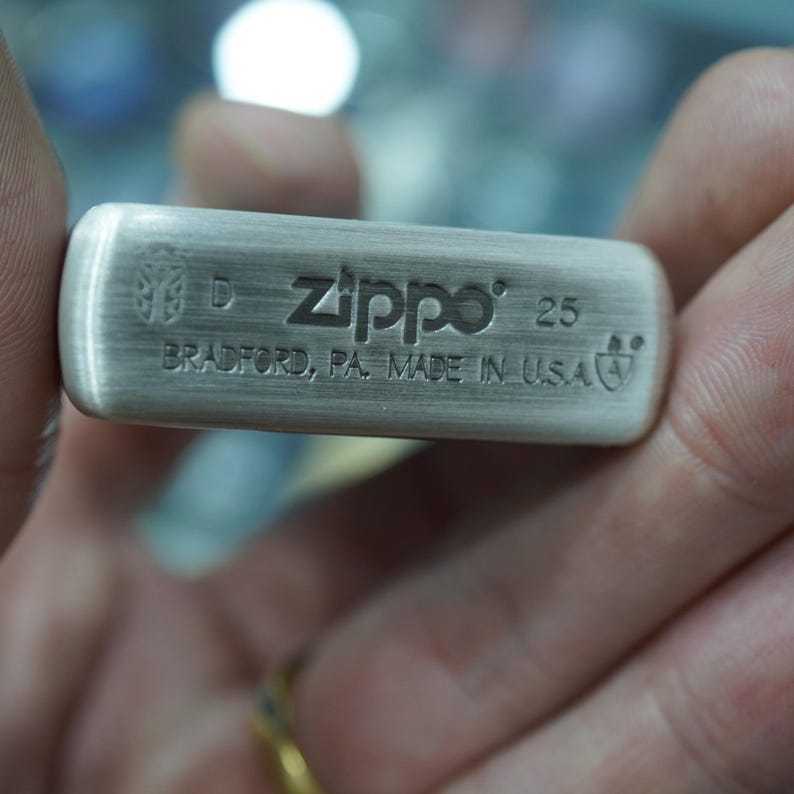 K&ouml;nnte beinhalten: Ein Feuerzeug der Marke Zippo aus geb&uuml;rstetem Metall mit dem Markennamen und der Gravur "25" auf der Oberfl&auml;che. Der Text "BRADFORD, PA. MADE IN U.S.A." ist ebenfalls sichtbar. Das Feuerzeug wird von einer Person gehalten.