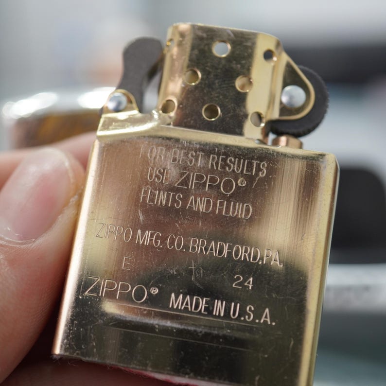 Może przedstawiać: Zbliżenie złotego zapalniczki Zippo. Zapalniczka ma napis "FOR BEST RESULTS USE ZIPPO FLINTS AND FLUID" oraz "ZIPPO MADE IN U.S.A." Zapalniczka jest prostokątna z zaokrąglonymi krawędziami i klapką na zawiasach.