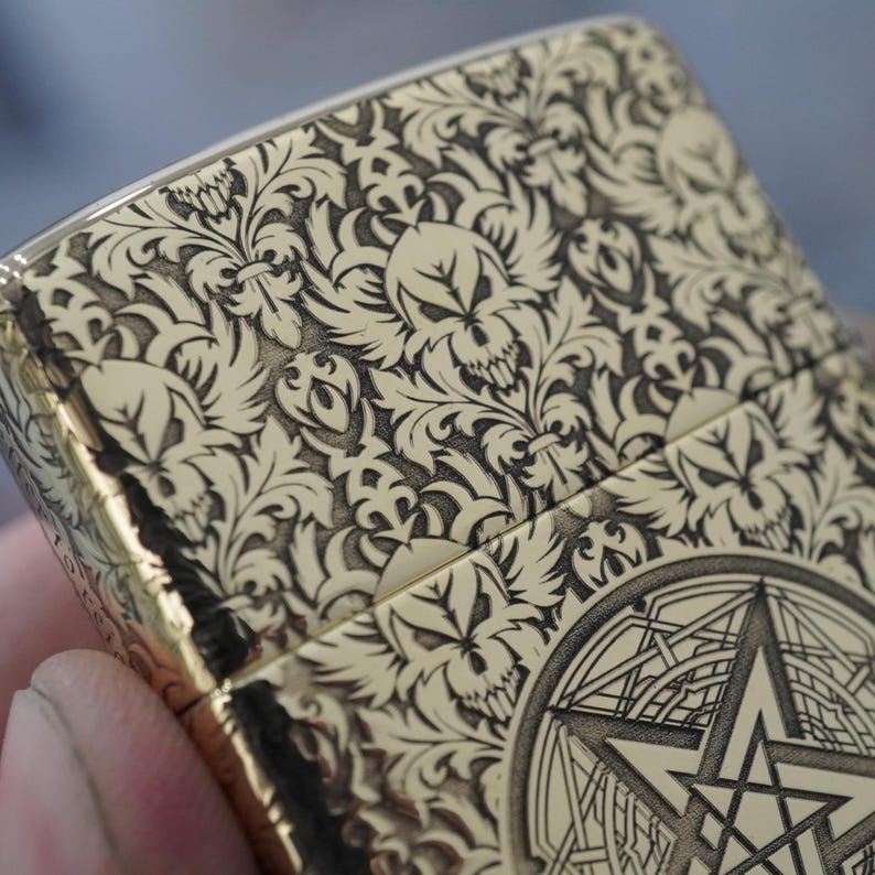 May include: A gold-coloured lighter with an intricate engraved design. The design features a repeating pattern of skulls and floral elements, with a geometric star emblem on the bottom. The lighter has a shiny, metallic finish.
