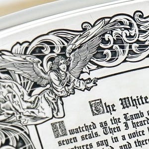 May include: Close-up of a silver-colored decorative plate with an engraved angel design. The angel has large wings and is surrounded by ornate scrollwork. Text on the plate reads "The White" and continues with additional text. The plate has a raised edge.