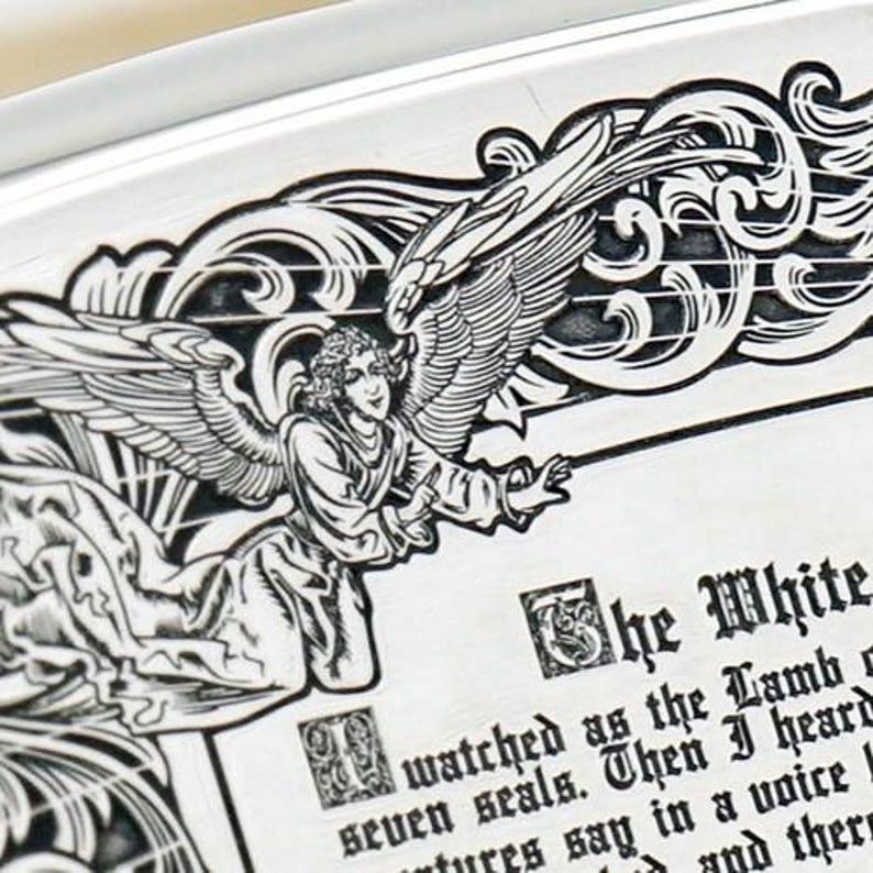 May include: Close-up of a silver-colored decorative plate with an engraved angel design. The angel has large wings and is surrounded by ornate scrollwork. Text on the plate reads "The White" and continues with additional text. The plate has a raised edge.