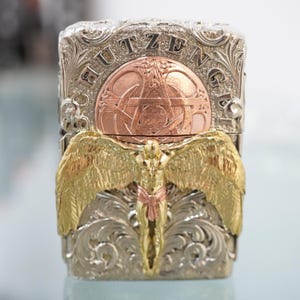 May include: Silver and gold-toned metal lighter with intricate engraved details. The lighter features a central copper-colored emblem with a star design and the name "HUTZENGER" engraved on the top. A gold-toned angel figure with outstretched wings is on the front.