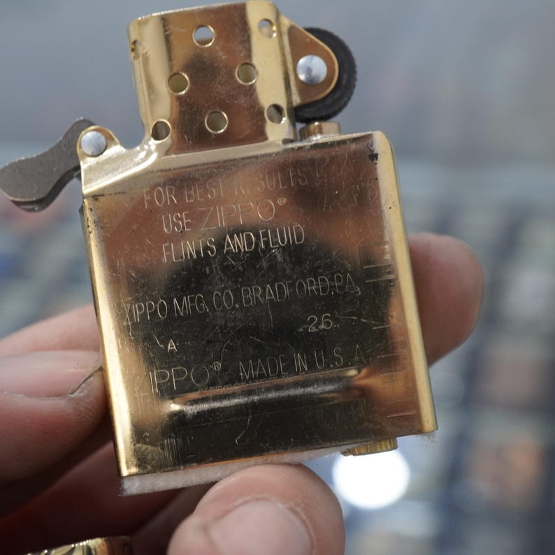 May include: A gold-coloured Zippo lighter, open to show the interior. The lighter has the text "FOR BEST RESULTS USE ZIPPO FLINTS AND FLUID" and "ZIPPO MFG. CO. BRADFORD, PA" printed on it. The lighter is made in the U.S.A.