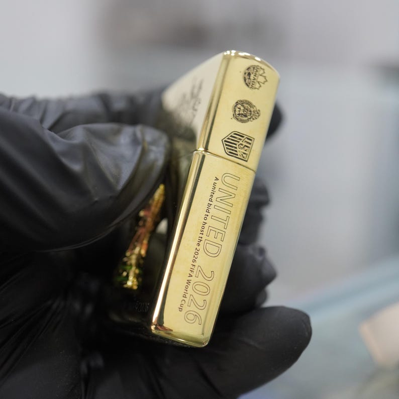 Puede incluir: Un encendedor de metal dorado con el texto grabado UNITED 2026 A united bid to host the 2026 FIFA World Cup. La superficie muestra tres peque&ntilde;os emblemas circulares y en forma de escudo que representan a Canad&aacute;, M&eacute;xico y Estados Unidos.