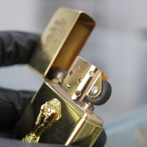 Puede incluir: Un encendedor de metal dorado con tapa abatible y acabado pulido. El encendedor presenta un emblema tridimensional dorado de un trofeo en la superficie frontal. La tapa est&aacute; abierta, revelando la chimenea interna y el mecanismo de encendido.