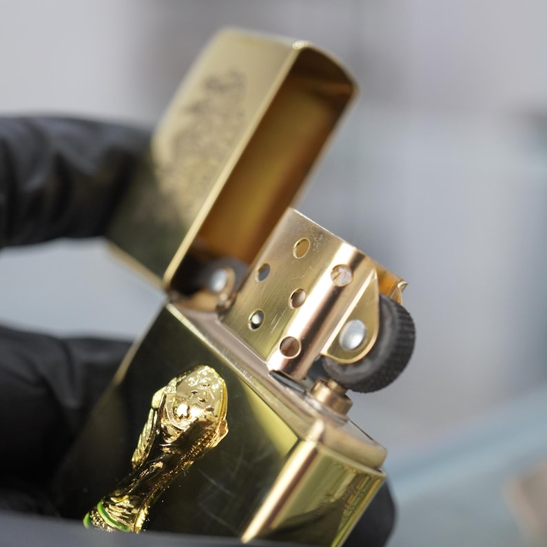 Puede incluir: Un encendedor de metal dorado con tapa abatible y acabado pulido. El encendedor presenta un emblema tridimensional dorado de un trofeo en la superficie frontal. La tapa est&aacute; abierta, revelando la chimenea interna y el mecanismo de encendido.