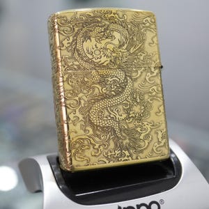 Pode incluir: Um isqueiro dourado com um desenho de drag&atilde;o gravado. O drag&atilde;o &eacute; intrincadamente detalhado, contra um fundo de nuvens rodopiantes. O isqueiro &eacute; retangular e tem uma tampa articulada. O isqueiro &eacute; exibido em um suporte prateado e preto.
