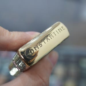 May include: A gold-coloured metal lighter with the word "CONSTANTINE" engraved on the side. The lighter has a rectangular shape with rounded edges and a decorative design near the top.