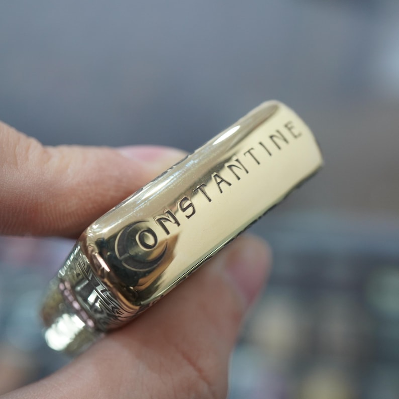 May include: A gold-coloured metal lighter with the word "CONSTANTINE" engraved on the side. The lighter has a rectangular shape with rounded edges and a decorative design near the top.