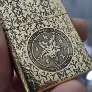 May include: A gold-coloured metal lighter featuring detailed engraved patterns. The design includes a central pentagram with a goat's head, surrounded by text. The lighter has a floral and skull motif.