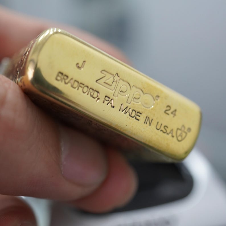 Pode incluir: Um close-up de um isqueiro Zippo dourado. O isqueiro &eacute; gravado com o nome da marca, a cidade de Bradford, PA, e "MADE IN U.S.A.". O isqueiro &eacute; segurado em uma m&atilde;o.