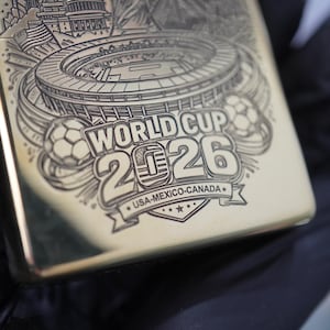 Puede incluir: Un encendedor de metal dorado con un grabado l&aacute;ser detallado de un estadio, balones de f&uacute;tbol y el texto WORLD CUP 2026 USA-MEXICO-CANADA. El grabado incluye un cero estilizado en el a&ntilde;o 2026 y una cinta decorativa con estrellas.