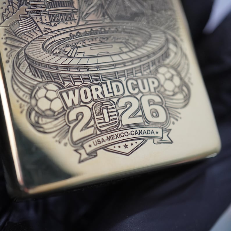 Puede incluir: Un encendedor de metal dorado con un grabado l&aacute;ser detallado de un estadio, balones de f&uacute;tbol y el texto WORLD CUP 2026 USA-MEXICO-CANADA. El grabado incluye un cero estilizado en el a&ntilde;o 2026 y una cinta decorativa con estrellas.