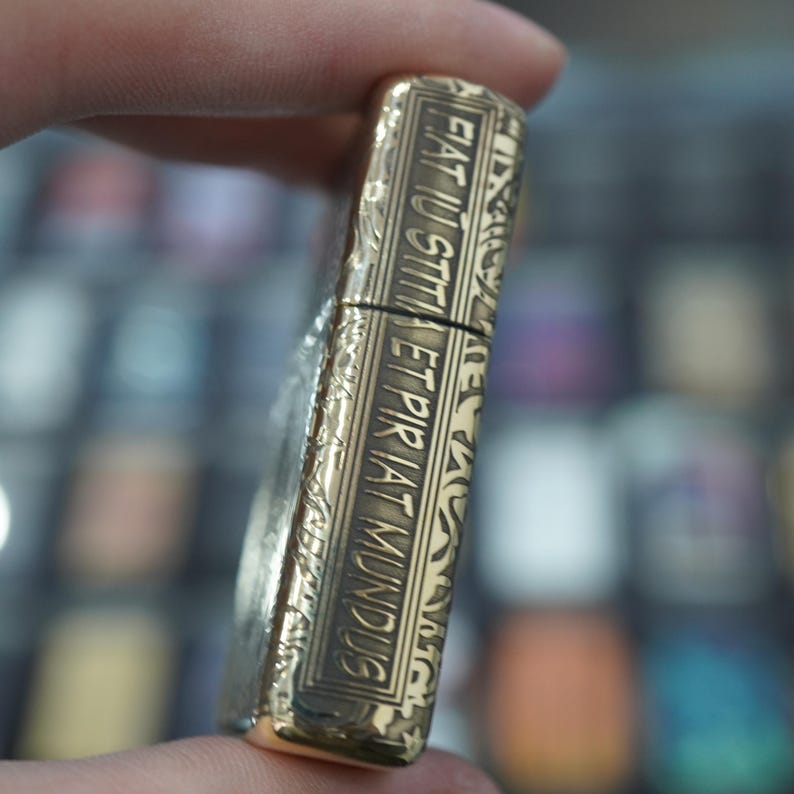 May include: A gold-coloured lighter with ornate engravings and the Latin phrase "FIAT IUSTITIA ET PIRIAT MUNDUS" is held in a hand. The lighter has a rectangular shape with a hinged lid and a textured surface.