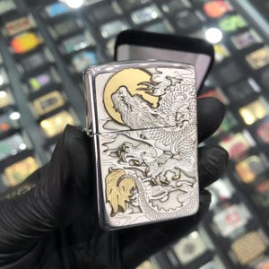 Może przedstawiać: Srebrna zapalniczka z szczeg&oacute;łowym wzorem smoka. Smok jest wytrawiony w srebrze ze złotymi akcentami, na tle złotego księżyca. Zapalniczka jest trzymana w czarnej rękawicy, z czarnym etui w tle.