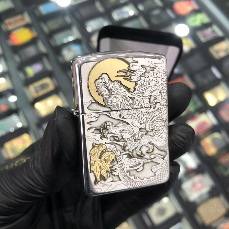 Może przedstawiać: Srebrna zapalniczka z szczeg&oacute;łowym wzorem smoka. Smok jest wytrawiony w srebrze ze złotymi akcentami, na tle złotego księżyca. Zapalniczka jest trzymana w czarnej rękawicy, z czarnym etui w tle.