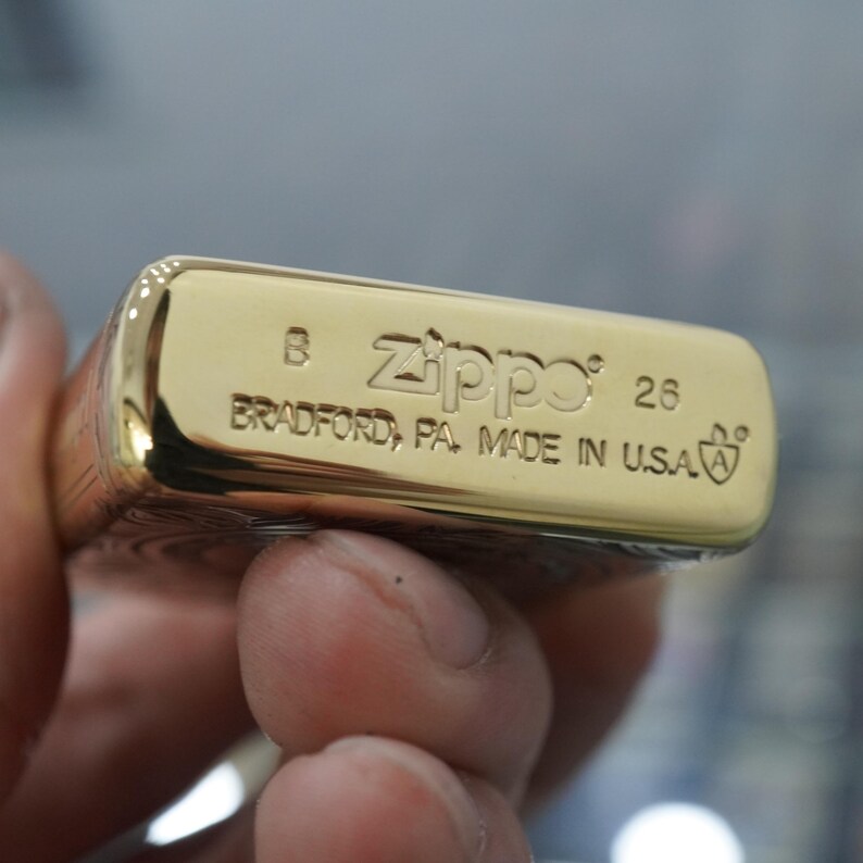May include: A gold Zippo lighter, with the text "Zippo 26" and "BRADFORD, PA. MADE IN U.S.A." engraved on the base. The lighter is held in a hand, with the focus on the engraved details and the shiny surface of the lighter.