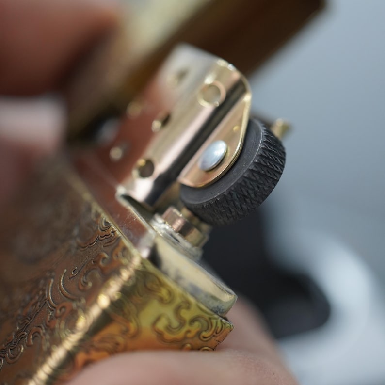 Pode incluir: Close-up de um isqueiro dourado com padr&otilde;es gravados intrincados. O mecanismo do isqueiro, incluindo a roda de pederneira e a dobradi&ccedil;a, &eacute; vis&iacute;vel. O isqueiro est&aacute; aberto, revelando seus componentes internos.