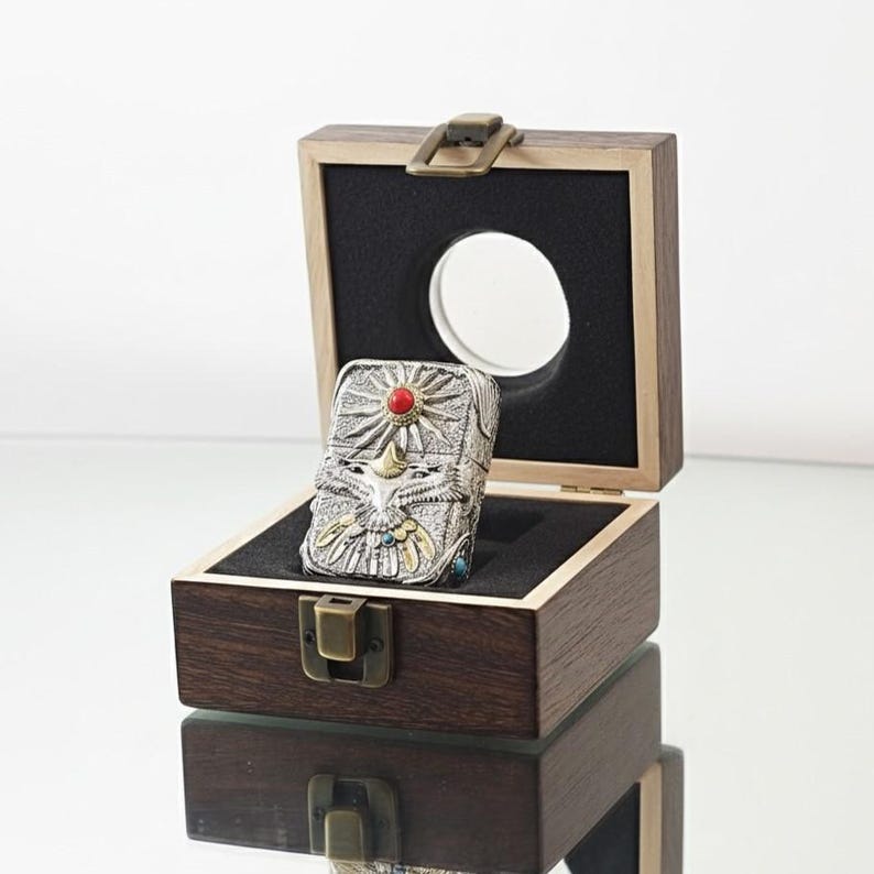 K&ouml;nnte beinhalten: Ein silberfarbener Ring mit einem kunstvollen Design, das einen Sonnenaufgang, einen Vogel und andere Details aufweist, besetzt mit einem roten Stein. Der Ring wird in einer offenen Holzkiste mit schwarzem Innenraum und rundem Ausschnitt pr&auml;sentiert.
