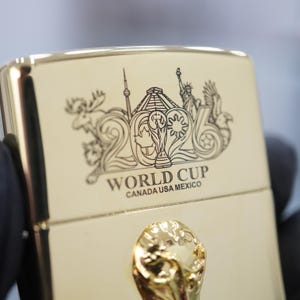 Puede incluir: Un encendedor de metal dorado con un grabado del n&uacute;mero 2026, el trofeo de la Copa Mundial y monumentos como la Estatua de la Libertad. Debajo del grabado, el texto dice WORLD CUP CANADA USA MEXICO. Se observa un emblema dorado en relieve.