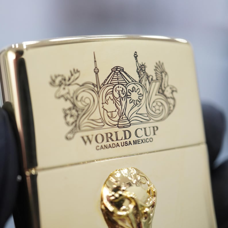 Puede incluir: Un encendedor de metal dorado con un grabado del n&uacute;mero 2026, el trofeo de la Copa Mundial y monumentos como la Estatua de la Libertad. Debajo del grabado, el texto dice WORLD CUP CANADA USA MEXICO. Se observa un emblema dorado en relieve.