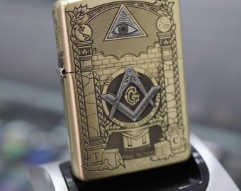 May include: A gold-colored lighter with intricate Masonic symbols engraved on its surface. The design includes an all-seeing eye within a triangle, a compass and square, and various architectural elements. The lighter is rectangular and has a hinged lid.