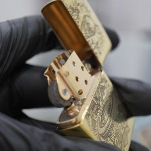 Puede incluir: Un encendedor de metal dorado con tapa abatible abierta, que revela la chimenea interna y el mecanismo de la rueda de pedernal. La superficie exterior presenta grabados detallados de l&iacute;neas negras, incluyendo el texto "WORLD" y "2020" en el lateral.
