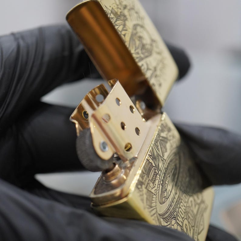 Puede incluir: Un encendedor de metal dorado con tapa abatible abierta, que revela la chimenea interna y el mecanismo de la rueda de pedernal. La superficie exterior presenta grabados detallados de l&iacute;neas negras, incluyendo el texto "WORLD" y "2020" en el lateral.
