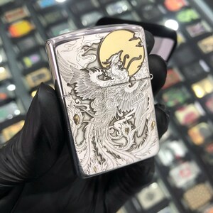 Może przedstawiać: Srebrna metalowa zapalniczka z grawerowanym wzorem feniksa. Zapalniczka posiada szczeg&oacute;łową ilustrację feniksa z pełnią księżyca w tle. Projekt jest w odcieniach srebra i złota.